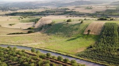 Aerial view of a picturesque countryside landscape with rolling hills, fields, trees, and a winding road. The tranquil beauty of nature is captured from above.