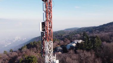 Telecommunication tower with antennas and satellite dishes on a mountain top overlooking a city in the valley below.