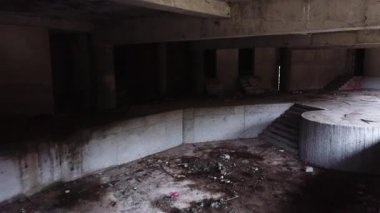 Empty swimming pool in an abandoned hotel building. Debris, concrete, and broken windows litter the space. Autumn woods visible outside.