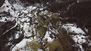 Aerial view of a remote village nestled in the snowy mountains. Sparsely populated, with winding roads and patches of green amidst the white landscape.