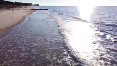 Low-angle aerial view of gentle waves lapping a sandy beach as the sun shines brightly over the sea, creating a glistening effect on the waters surface.