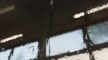 Water droplets slide down the side window at a car wash, glistening under the bright afternoon sun. The view includes reflections and a serene ambiance in this auto detailing environment.