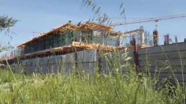 Construction work is underway on a new structure surrounded by wild grass and fencing. Cranes are visible as workers manage the site under a bright sky.