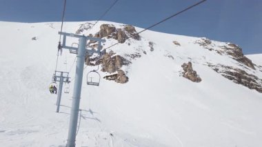 A chairlift ascends through a stunning winter landscape filled with snow and rocky peaks. The bright blue sky enhances the beauty of this pristine mountain environment.