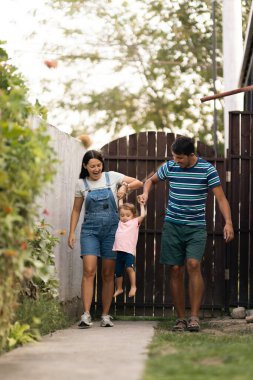 A cheerful family enjoys a walk outdoors in their garden, lifting their child playfully. A moment reflecting happiness, familial bond, and leisure in nature. Bright and full of connection.