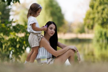 A tender scene of a child combing her mothers hair, reflecting family love and bonding moments in a calm natural landscape with trees and water.