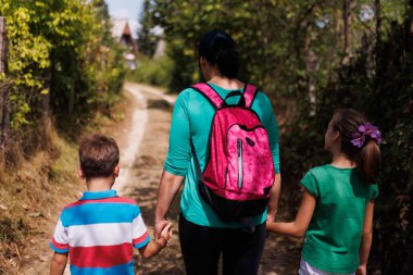 A woman with a pink backpack walks with two children on a sunlit dirt road surrounded by green trees, creating a sense of family bond and outdoor activity.