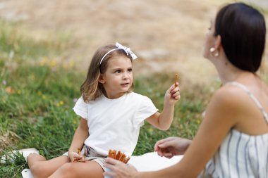 A young mother and her daughter sharing snacks during a peaceful picnic on a sunny day in a natural setting.