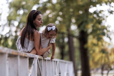 A mother and her young daughter, engaged in a tender moment, enjoy a peaceful day in a lush, green park environment, featuring expressions of care, connection, and natural beauty.