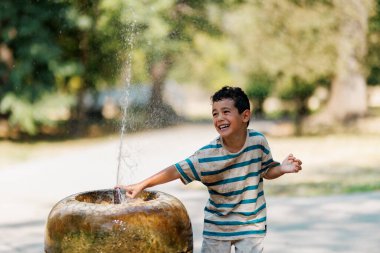 A joyful young boy interacts with a fountain, splashing water on a bright summer afternoon amidst greenery, showcasing childhood happiness, carefree play, and outdoor fun.