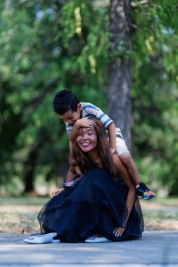 A cheerful young woman kneeling on the ground in a park as a boy holds onto her shoulders from behind, both enjoying a joyful moment together.
