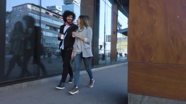 Happy young couple walking casually through a vibrant city, enjoying coffee and conversation.