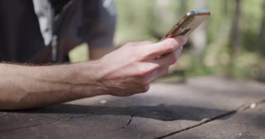Relaxing in a peaceful outdoor environment, an individual engages with their smartphone while seated on a rustic wooden table. The vibrant greenery enhances the tranquil atmosphere.