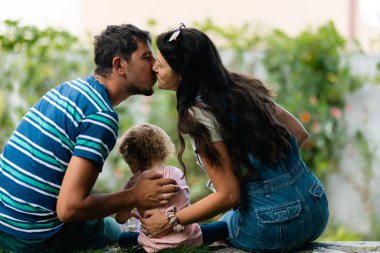 A warm family moment featuring parents embracing and sharing a kiss in a natural setting, with their child in between, evoking a sense of love, connection, and togetherness.
