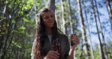 A young woman is immersed in a lush forest, engaged in outdoor activities. She holds a tool and expresses her passion for nature while surrounded by tall trees and greenery.