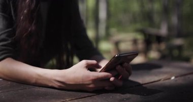 Enjoying a peaceful moment outdoors at a picnic table, a person engages with their smartphone in a lush forest. The sun shines brightly, enhancing the serene atmosphere.
