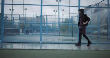 A young woman wearing a hoodie walks thoughtfully across a sports court, surrounded by a blue fence, on an overcast day, conveying a sense of solitude and introspection.