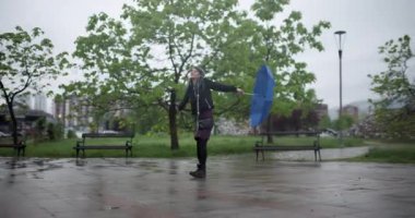 A young woman enjoys the rainy weather by dancing joyfully in a park, holding a large blue umbrella.