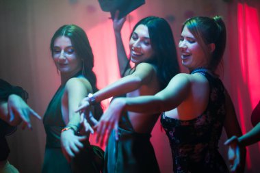 Three young women express lively energy as they enjoy dancing under vibrant light effects, exhibiting happiness and camaraderie at a celebration.