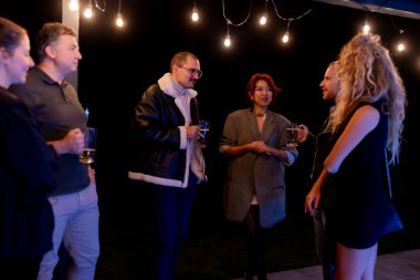 A small group of friends enjoying casual conversations and drinks together outdoors in a nighttime setting decorated with string lights, exemplifying friendship, leisure, and relaxed moments.