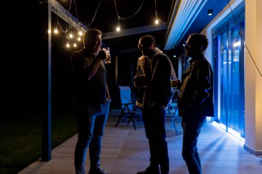 Three friends drink together in a well-lit patio area during night, enjoying a relaxed atmosphere with string lights enhancing the ambiance.