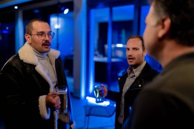 Three people engage in conversation at a cozy evening gathering, enjoying beverages in a relaxed setting illuminated by blue ambient lighting