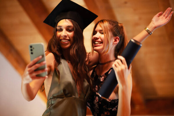 Two friends joyfully celebrate a graduation achievement. One person wears a cap, and the pair captures the moment together with a phone selfie, symbolizing friendship, achievement, and cherished