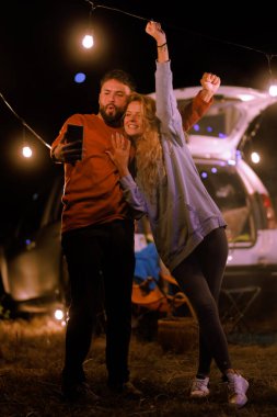 Two friends are celebrating at night under glowing string lights. They stand near a car, sharing drinks and laughter, enjoying the warmth of the evening and each others company.