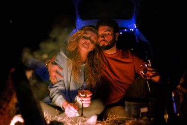 A couple sits together beside a campfire, surrounded by nature at night. They are warmly dressed, holding glasses and enjoying the moment, creating a romantic atmosphere.