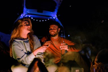 Two friends relax by a campfire at night, sharing stories and laughter. The glowing fire illuminates their smiling faces as they hold drinks, creating a warm and friendly atmosphere.