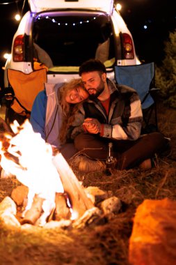 A couple sits close to a warm campfire, surrounded by nature. They share a moment of affection, wrapped in blankets while the night sky sparkles with stars.