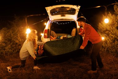 A couple is preparing for a night outdoors, laying out a blanket next to their vehicle. String lights illuminate the scene, creating a warm and inviting atmosphere in nature.