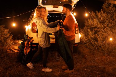 A couple is happily preparing a tent together under warm string lights near their vehicle. The scene is set in a peaceful outdoor location at night, surrounded by nature.