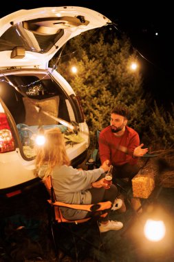 Two people sit by their car, enjoying drinks and conversation at night. String lights provide a warm glow in the forest, creating a cozy atmosphere.