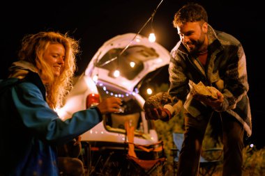 Two friends are gathered by their car at night, smiling and sharing food. Twinkling lights add to the warm ambiance while they enjoy their time outdoors.