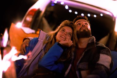 Two individuals sit closely together under a starry sky, gazing up and sharing a warm moment by a fire, surrounded by the inviting glow of fairy lights.