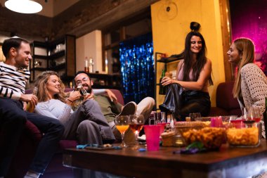 Five friends gather in a relaxed living room setting, enjoying a fun party with food, drinks, and laughter, sharing a joyful and social evening.