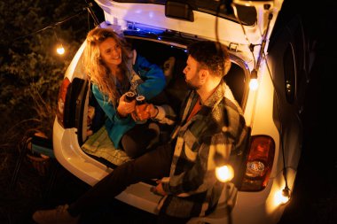 Two people share drinks while sitting in the back of a car adorned with string lights. It is night, and the atmosphere is warm and inviting, surrounded by nature.