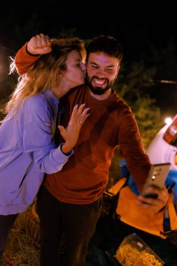 A couple is having a joyful moment while camping at night. They are smiling, taking a selfie, and celebrating together in a serene outdoor setting surrounded by nature.