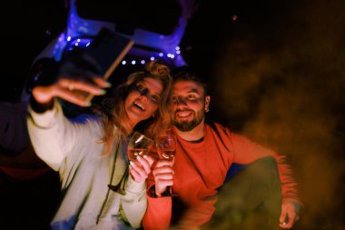 A couple smiles and poses for selfies while holding wine glasses beside a warm campfire. The night is cool, and twinkling lights add charm to the scene.