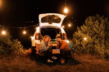 A couple sits on camping chairs next to their parked car in a field, illuminated by warm fairy lights. They enjoy snacks and drinks while chatting happily in the evening.