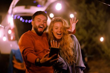 A couple is happily engaging in a video call while camping at night. They are surrounded by string lights, with a cozy atmosphere that enhances their joyful moment together.