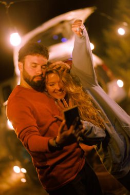 Two people are having fun together during a nighttime camping trip. They are smiling and taking a selfie with string lights creating a warm, inviting glow around them.