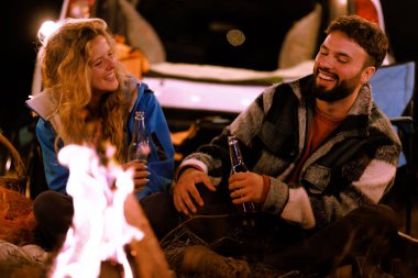 Two friends sit by a campfire at night, laughing together as they enjoy drinks and the warmth of the flames. The scene is relaxed and inviting, set in nature.