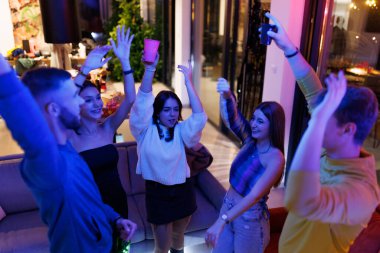A lively gathering of friends celebrating with fun and dance inside a well-lit and colorful indoor setting. Everyone appears joyful and engaged in the festive atmosphere.