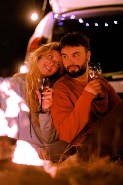 A couple sits close together by a campfire, holding glasses. They enjoy their time under the stars, sharing laughter and warmth in a peaceful outdoor atmosphere.