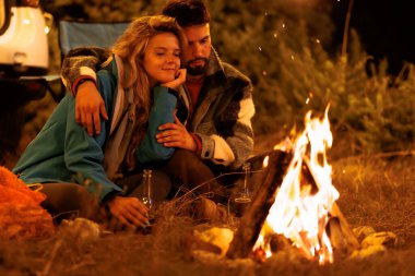 A couple sits closely by a campfire at night, enjoying each others company. They are cuddled together, surrounded by nature, with drinks in hand, creating a warm and intimate atmosphere.
