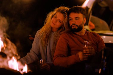 Two friends relax by a warm campfire, holding drinks and enjoying each others company. The scene is serene with dark surroundings and the glow of the fire illuminating their faces.