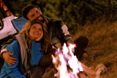 A couple relaxes by a crackling campfire, sharing smiles and warmth. Surrounded by trees and their vehicle, they enjoy a peaceful evening in the woods, creating memories together.