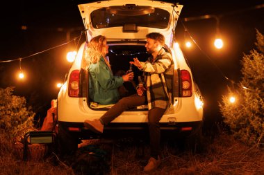 Two people are seated in the back of a car, surrounded by soft glow of string lights. They are sharing drinks and smiles in a serene nighttime outdoor environment, creating a cozy vibe.